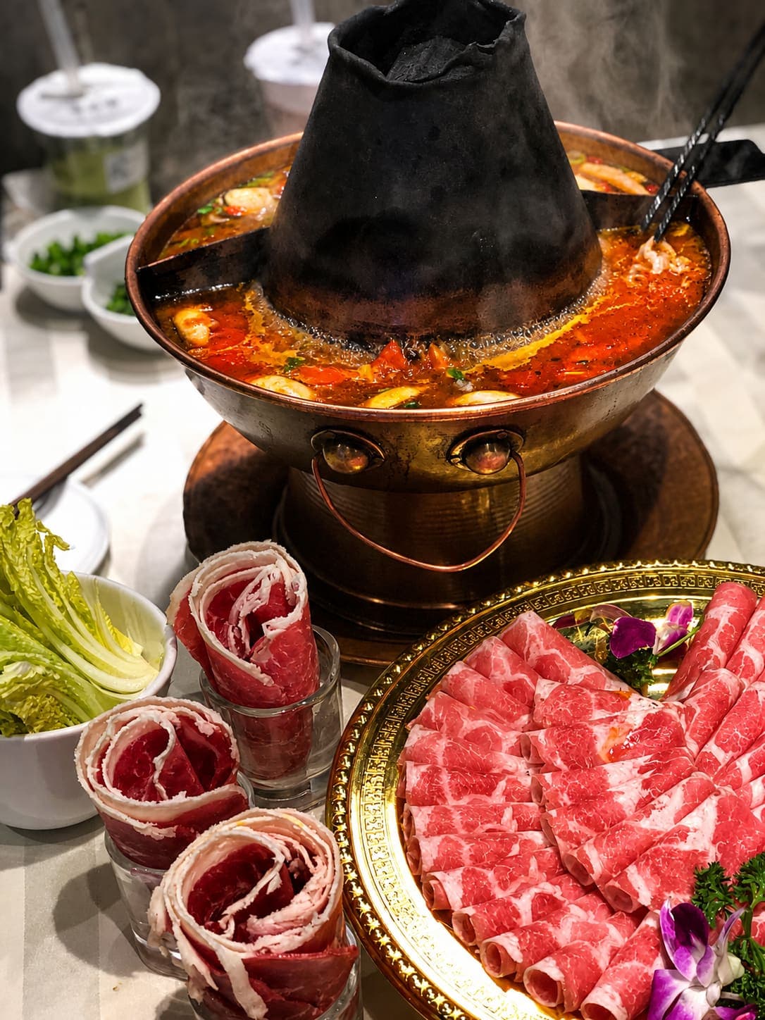 A copper Beijing hotpot with its chimney smoking, sliced lamb shoulder and sesame dipping sauce around it
