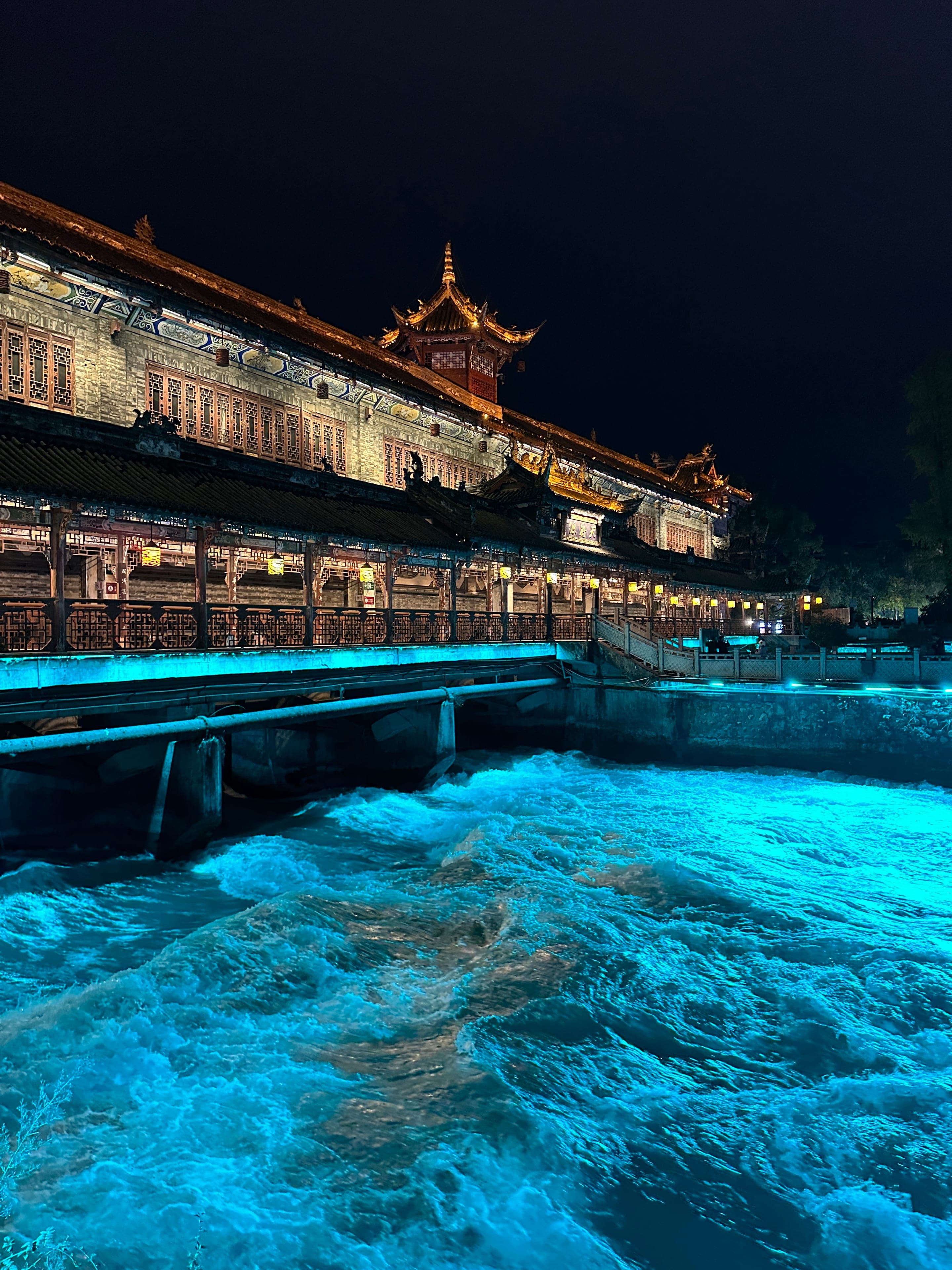 A night view of the Dujiangyan inner channel with the river water lit from beneath in luminous deep blue, the 2,000-year-old engineering glowing under the surface