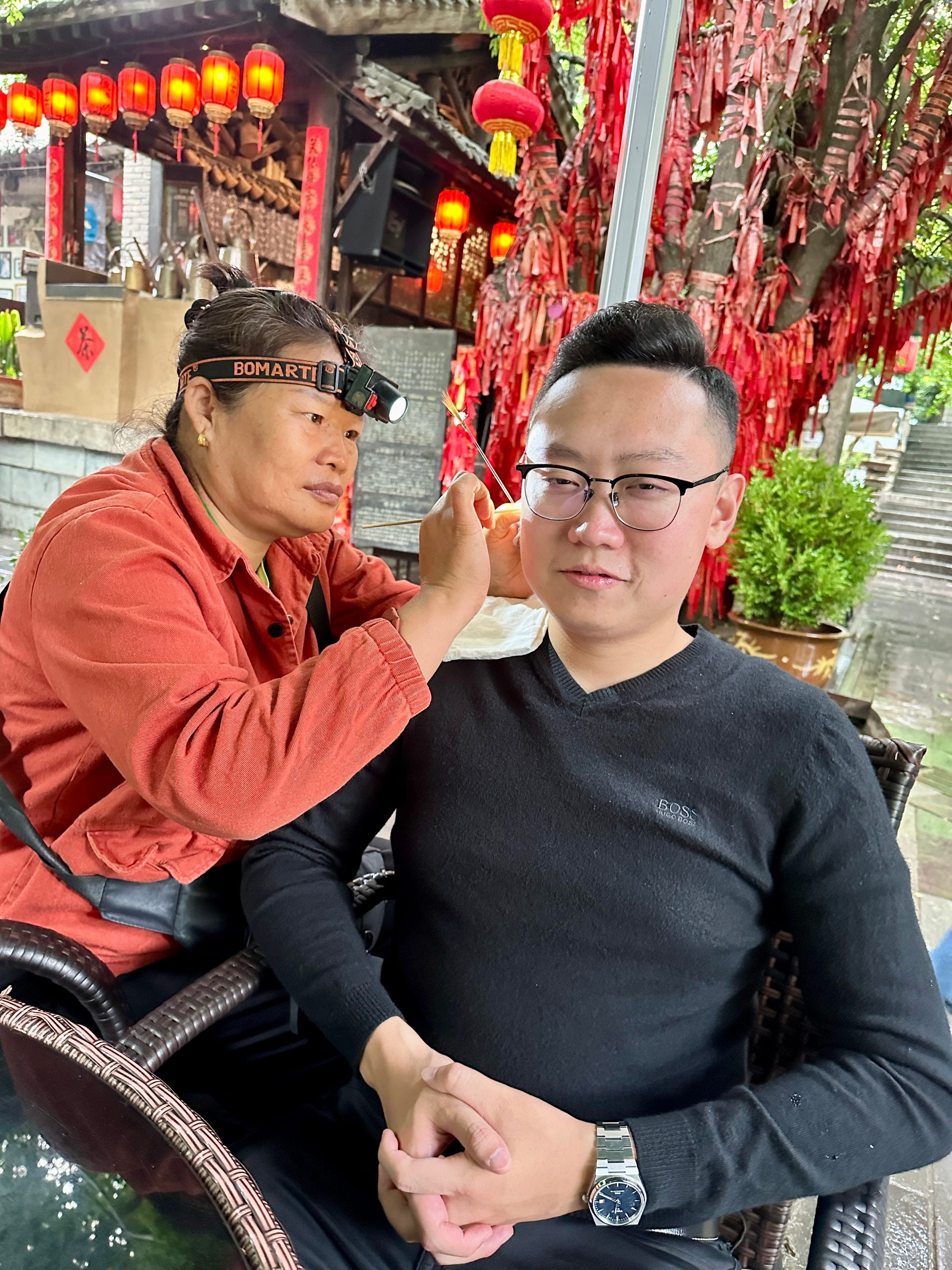 A Sichuan ear-cleaning master with a headlamp working on a seated customer at an outdoor teahouse, red lanterns hanging behind