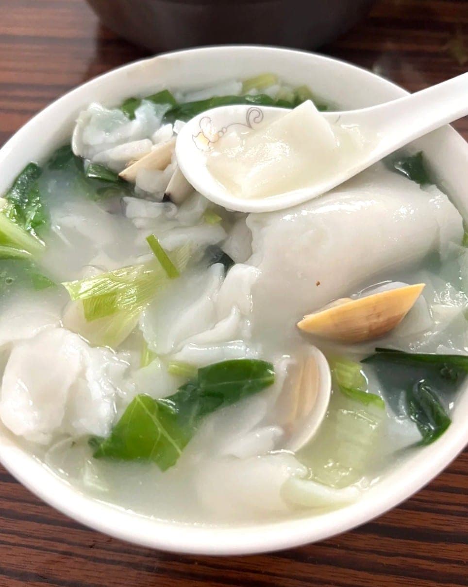 A bowl of Fuzhou guo bian hu, white rice-paste ribbons and a clam in a clear broth, spoon lifting one piece
