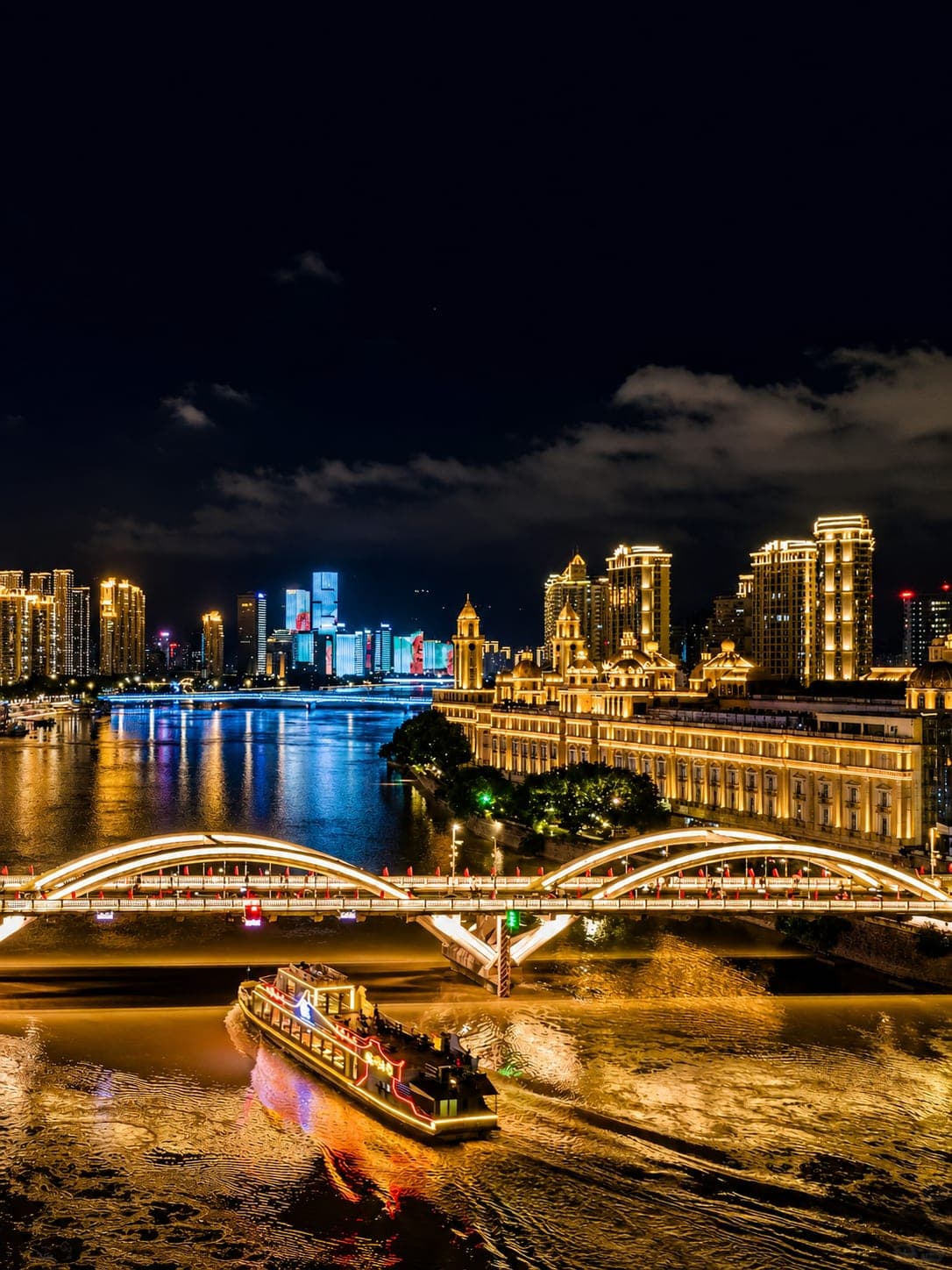 A lit cruise boat passing under a bridge on the Min River at night, with Zhongzhou Island's European-style block and the Fuzhou skyline behind