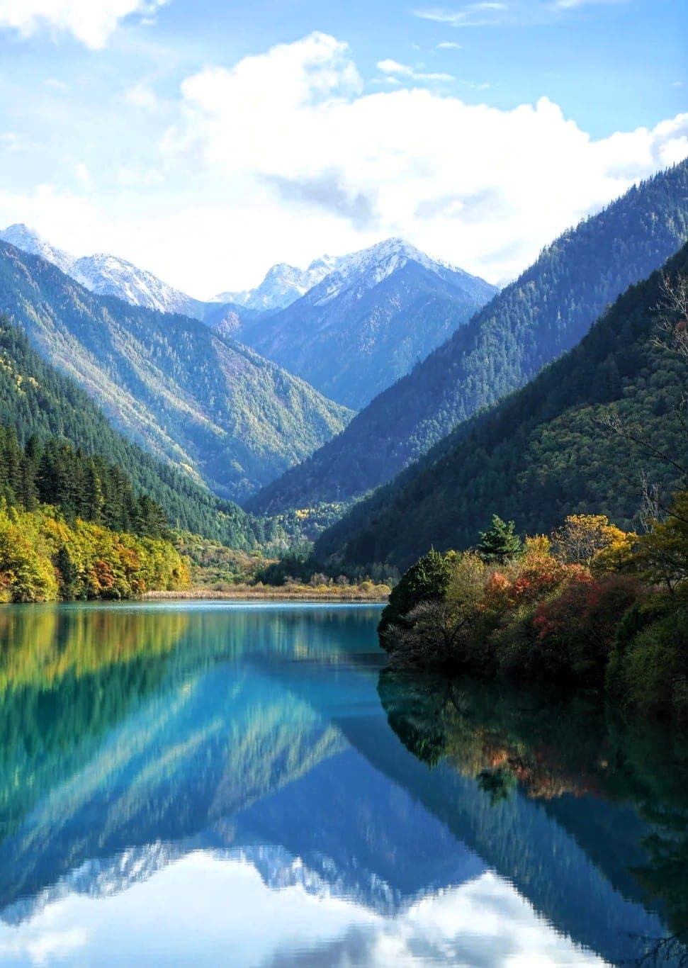 Rhinoceros Lake at Jiuzhaigou in autumn, a wide sheet of still blue and jade water holding the reflection of snow-capped peaks and the forested valley walls, with gold and russet foliage along both shores