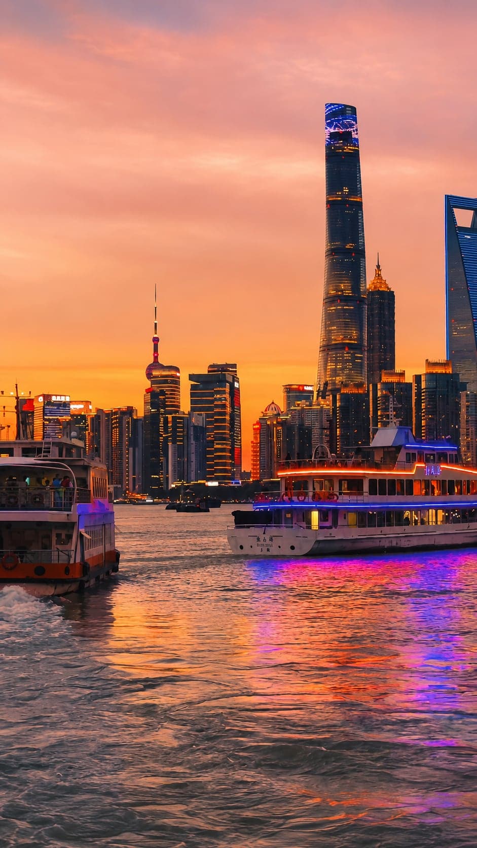 Two Pujiang ferries crossing the Huangpu at sunset, the Pudong skyline of Shanghai Tower, Jin Mao, Shanghai World Financial Center and the Oriental Pearl glowing against an orange sky