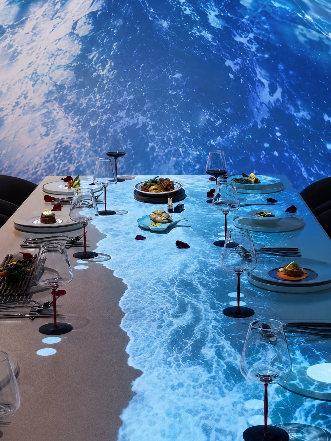 A long communal dining table set for fine dining, plated courses arranged along its centre, the entire table surface and back wall layered with a high-resolution sea projection, the projected waves rolling onto the table itself, a Shanghai holographic dinner