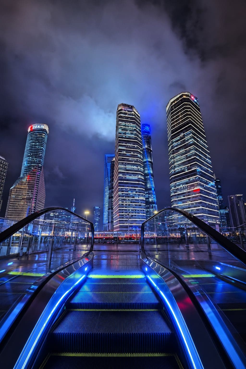 The Lujiazui pedestrian skybridge at night, blue LED runners on the escalator leading up to Pudong's supertall glass towers above the Huangpu
