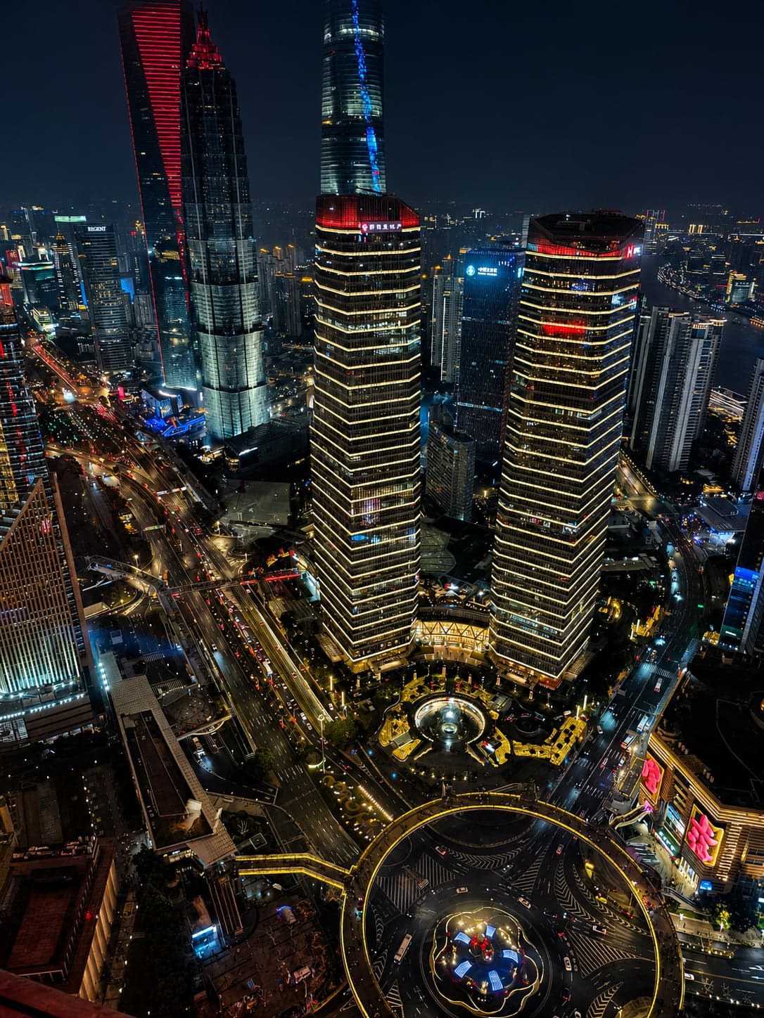Night aerial view down onto Lujiazui from the Shanghai Tower observation deck, lit twin office towers and the roundabout traffic circle below, the city stretching out in deep dark