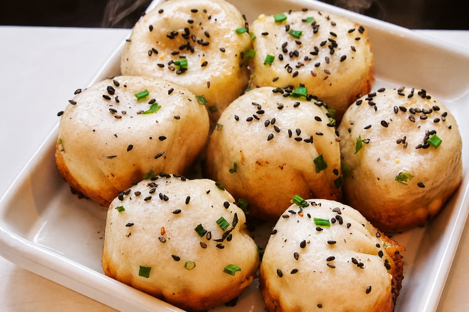 Six Shanghai shengjianbao on a white enamel plate at a city counter, sesame and scallion dusted across the soft tops, the crisp golden bases just visible at the edges