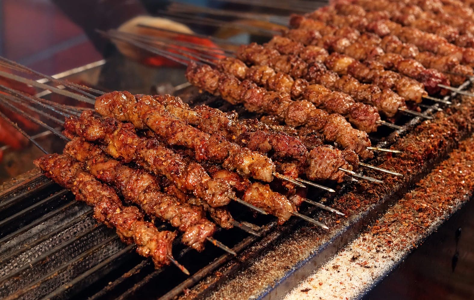 Xi'an lamb skewers grilling over charcoal, with raw lamb pieces ready at the back and cumin and chilli powder scattered along the rail