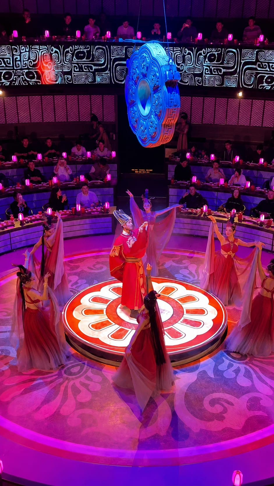 A soloist in red Tang-court robe at the centre of a circular stage, ringed by dancers in pastel gauze, with banquet tables curving around the audience behind, Xi'an