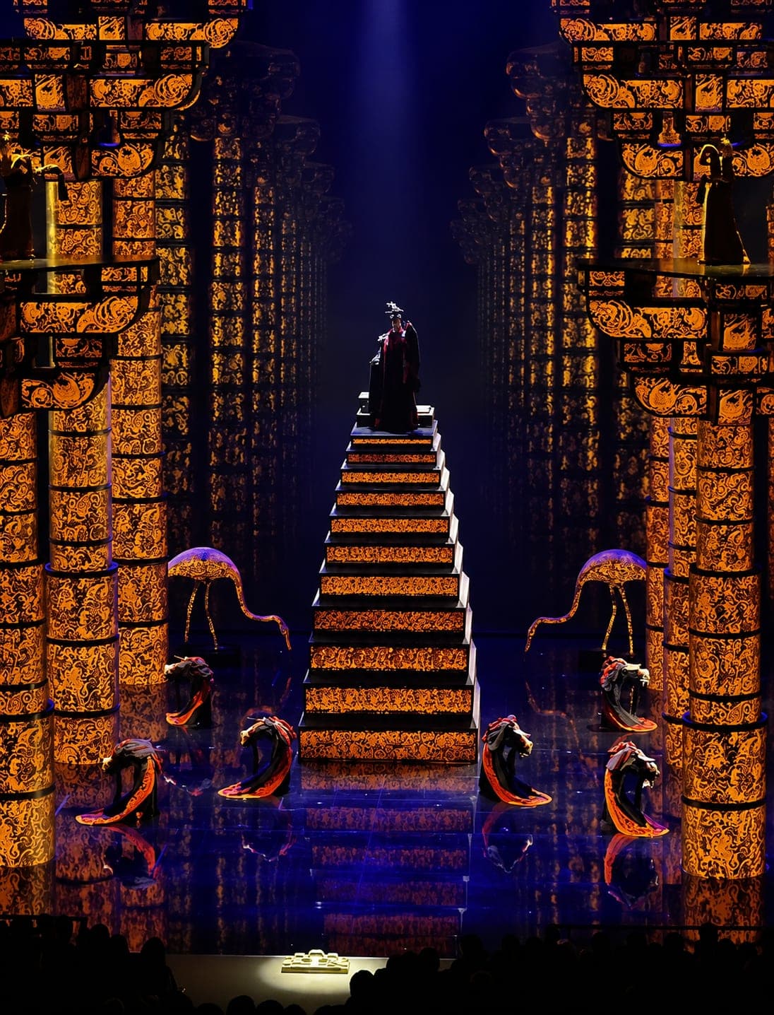 A robed figure at the apex of a central staircase, flanked by symmetrical Qin-style palace walls in orange relief beneath blue stage light, the Great Qin show, Xi'an