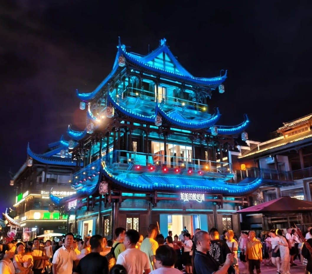 夜色中的大庸古城，一座层叠飞檐的木楼通体点亮蓝光，楼下街市游人如潮