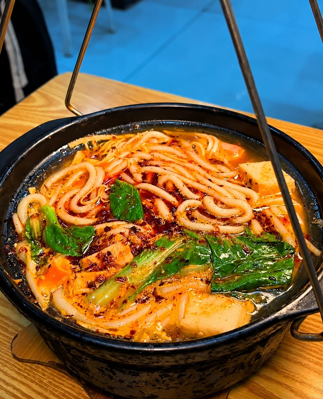 Kunming crossing-bridge noodles, a deep blue-and-white broth bowl ringed by side plates of sliced meats, quail egg, chrysanthemum leaves and rice noodles for table assembly