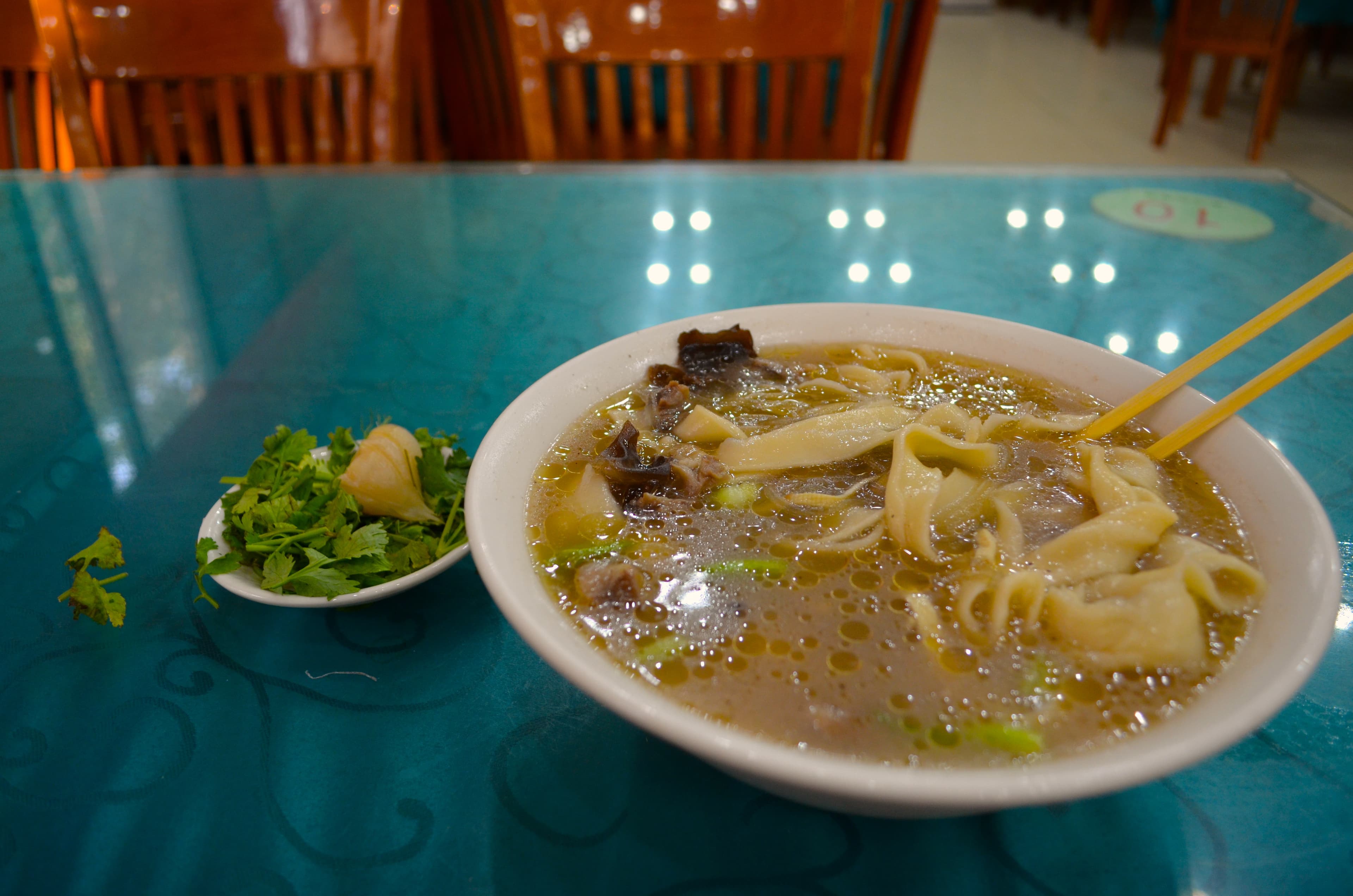 A large ceramic bowl of Zhengzhou Hui Mian, wide hand-pulled noodles in lamb broth with lily buds, wood ear and bean threads